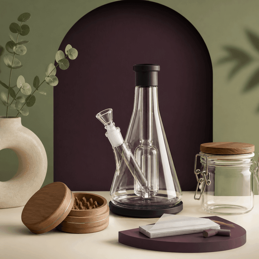 Bong transparente junto a un grinder abierto, un tarro vacío con cierre metálico, papeles de liar y boquillas sobre una mesa. Bong transparente junto a un grinder abierto, un tarro vacío con cierre metálico, papeles de liar y boquillas sobre una mesa.