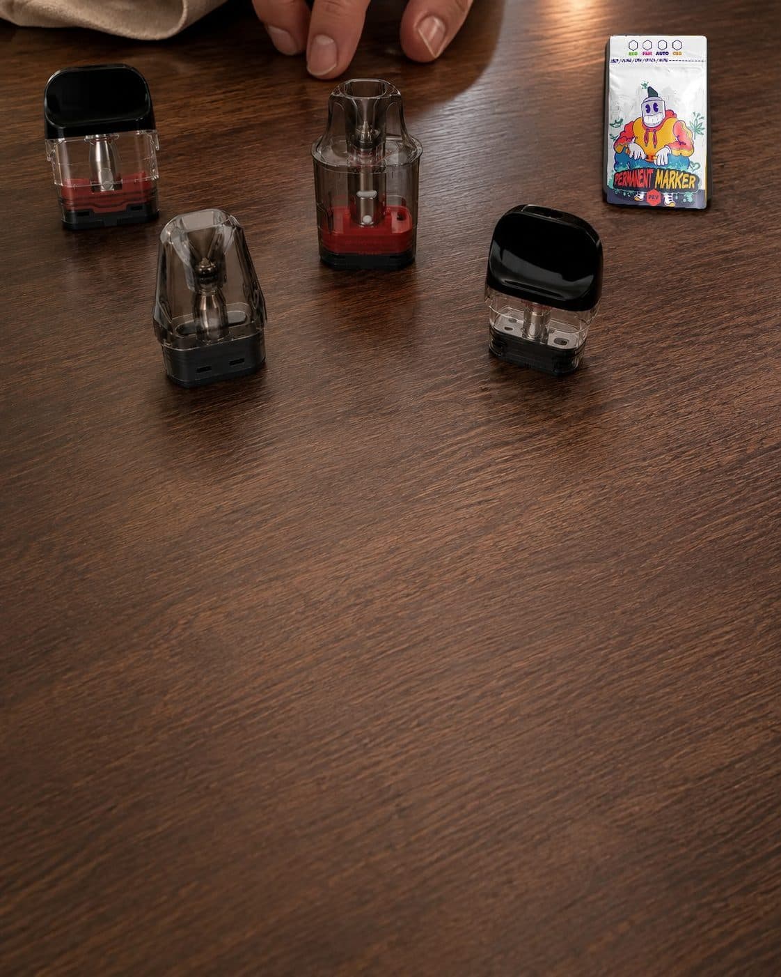 Cuatro cartuchos de vapeo de diferentes formas sobre una mesa de madera, junto a un sobre con el texto «Permanent Marker». Cuatro cartuchos de vapeo de diferentes formas sobre una mesa de madera, junto a un sobre con el texto «Permanent Marker».