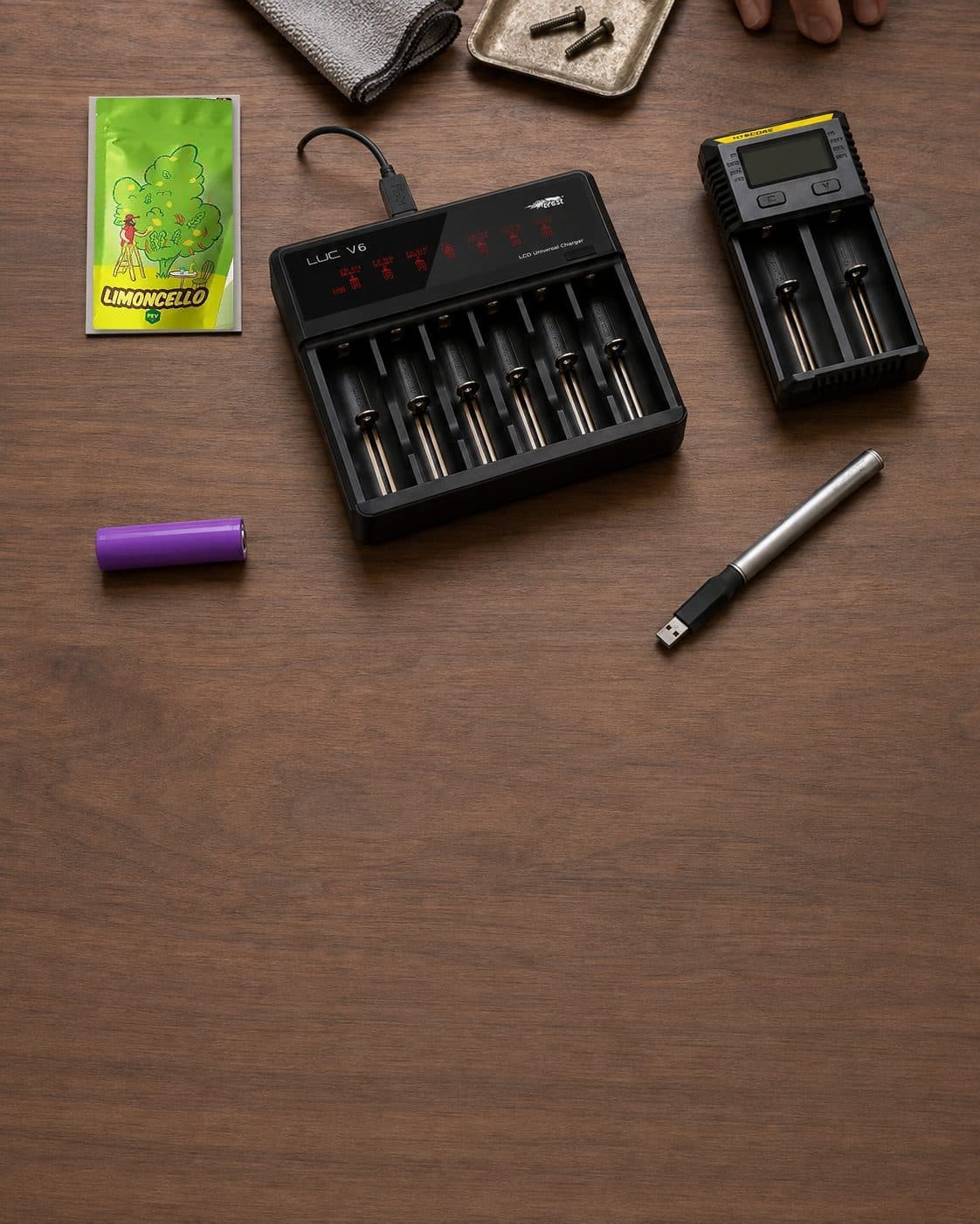 Dos cargadores de baterías de seis y dos ranuras, una batería morada y un dispositivo alargado con conector USB sobre una mesa. Dos cargadores de baterías de seis y dos ranuras, una batería morada y un dispositivo alargado con conector USB sobre una mesa.