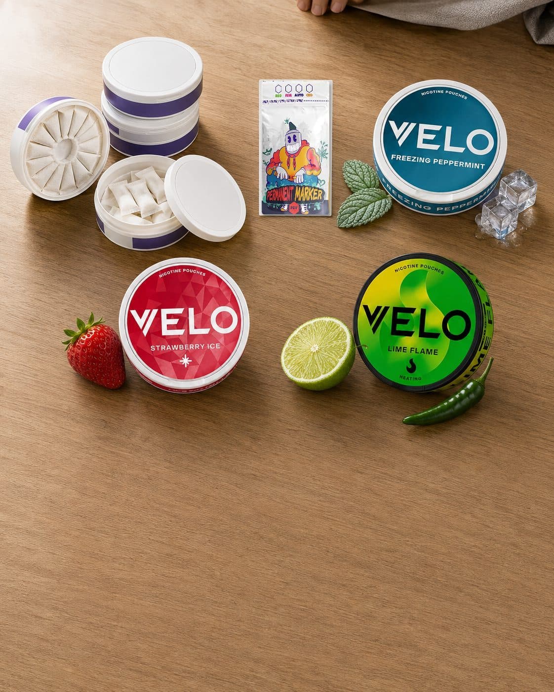 Latas VELO de menta, fresa y lima junto a recipientes con bolsitas blancas, una fresa, media lima y una guindilla sobre madera. Latas VELO de menta, fresa y lima junto a recipientes con bolsitas blancas, una fresa, media lima y una guindilla sobre madera.
