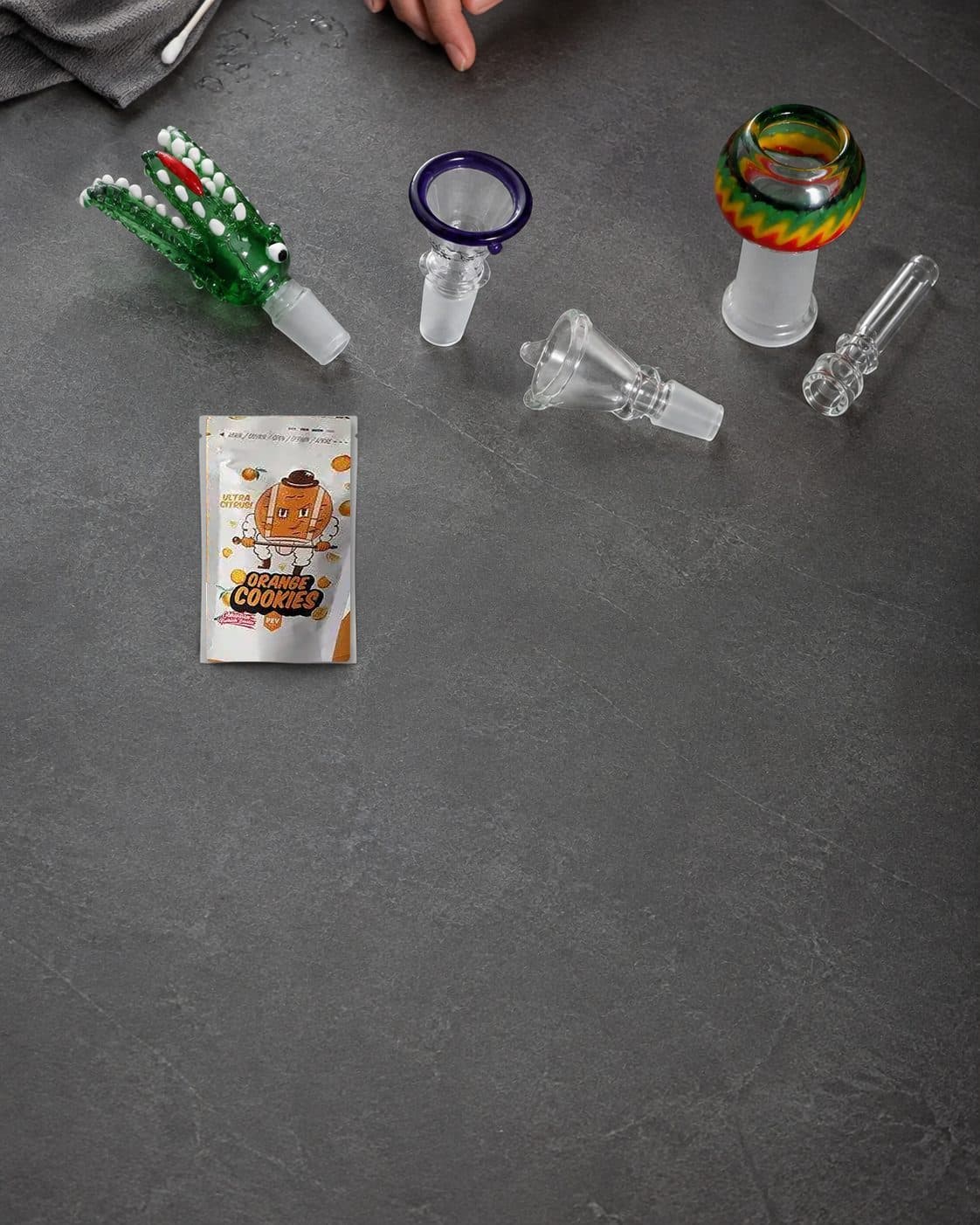 Cazoletas de cristal con figura verde dentada, borde violeta y diseño multicolor, junto a accesorios transparentes y un sobre Orange Cookies. Cazoletas de cristal con figura verde dentada, borde violeta y diseño multicolor, junto a accesorios transparentes y un sobre Orange Cookies.