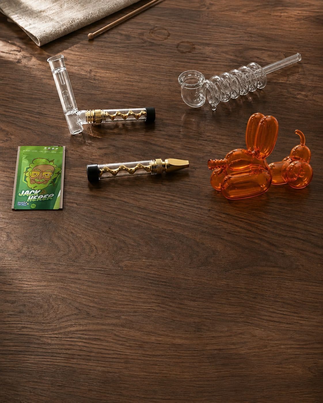 Pipas de distintos diseños sobre madera: una transparente en espiral, otra naranja con forma de perro globo y piezas con detalles dorados. Pipas de distintos diseños sobre madera: una transparente en espiral, otra naranja con forma de perro globo y piezas con detalles dorados.
