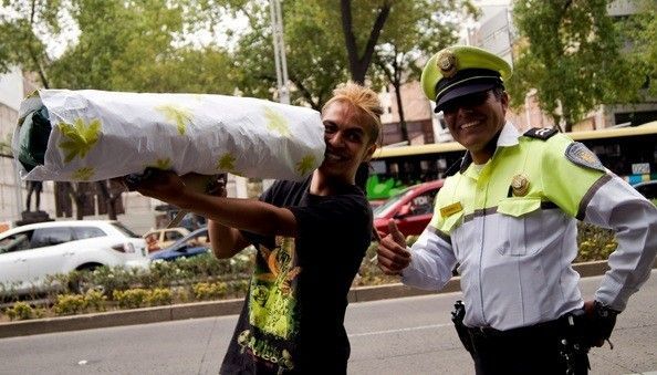 Una persona sostiene un gran objeto con forma de cigarrillo y hojas de cannabis junto a un agente uniformado que levanta el pulgar en la calle. Una persona sostiene un gran objeto con forma de cigarrillo y hojas de cannabis junto a un agente uniformado que levanta el pulgar en la calle.