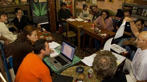Varias personas reunidas alrededor de mesas con documentos y un portátil; una sostiene una larga tira de papel.