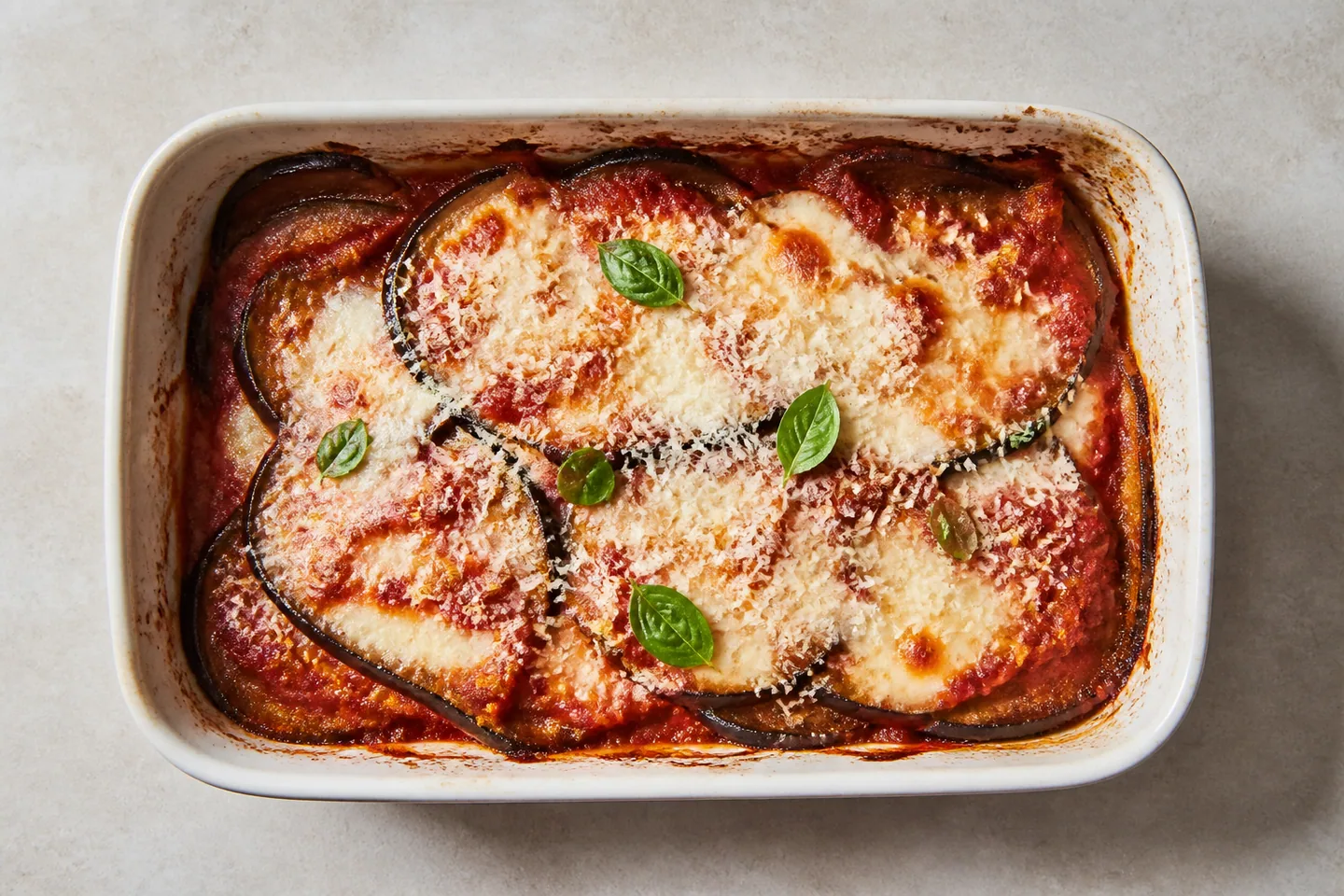 Vista cenital de berenjenas al horno en una fuente rectangular blanca, con salsa de tomate, queso dorado y hojas de albahaca. Vista cenital de berenjenas al horno en una fuente rectangular blanca, con salsa de tomate, queso dorado y hojas de albahaca.