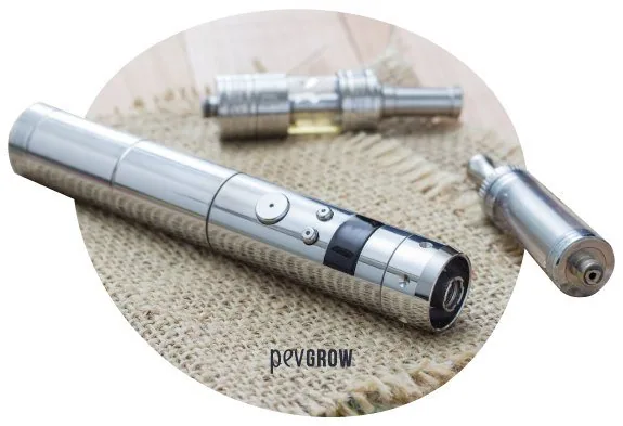 Vaporizador cilíndrico plateado con botones y pantalla, junto a dos piezas separadas sobre un tejido beige.