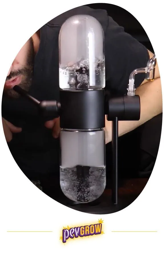Bong giratorio con dos cámaras transparentes, agua y burbujas, cuerpo central negro y boquilla curva lateral, junto a una persona. Bong giratorio con dos cámaras transparentes, agua y burbujas, cuerpo central negro y boquilla curva lateral, junto a una persona.