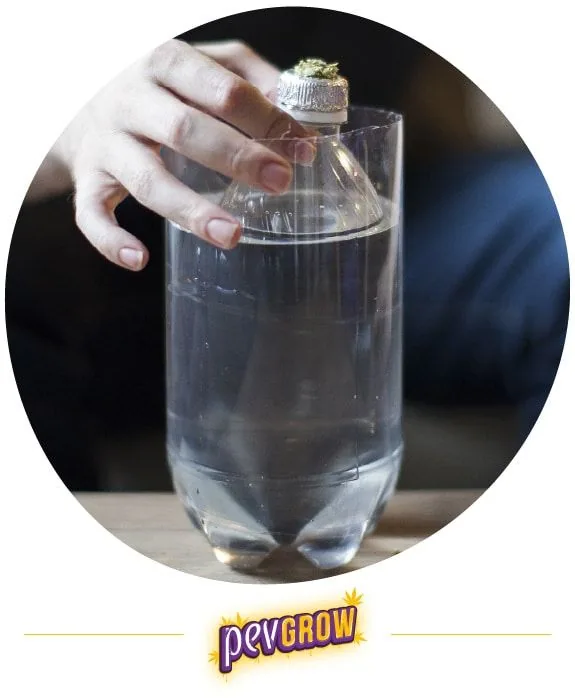 Una mano sujeta una botella con cazoleta de aluminio en el cuello, dentro de otro recipiente de plástico con agua, como bong casero. Una mano sujeta una botella con cazoleta de aluminio en el cuello, dentro de otro recipiente de plástico con agua, como bong casero.