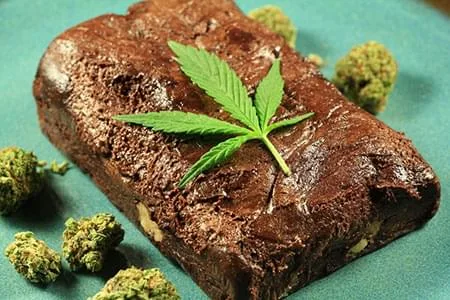 Brownie de chocolate rectangular con una hoja de cannabis encima, rodeado de cogollos sobre una superficie turquesa. Brownie de chocolate rectangular con una hoja de cannabis encima, rodeado de cogollos sobre una superficie turquesa.