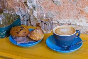 Taza azul de café con espuma, dos piezas de repostería y un vaso de agua sobre una mesa amarilla junto a una pared de ladrillo. Taza azul de café con espuma, dos piezas de repostería y un vaso de agua sobre una mesa amarilla junto a una pared de ladrillo.