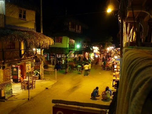 Calle nocturna con locales iluminados, terrazas y peatones, vista desde un balcón con dos personas sentadas junto a la acera.