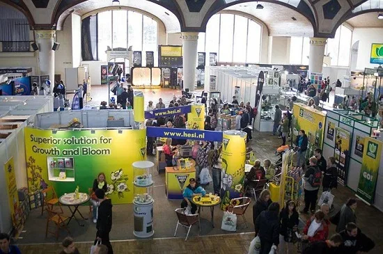 Vista elevada de una feria con visitantes y puestos de cultivo, en un pabellón con grandes ventanales y arcos. Vista elevada de una feria con visitantes y puestos de cultivo, en un pabellón con grandes ventanales y arcos.