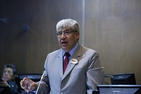 Hombre de pelo canoso y gafas, con traje gris y corbata roja, hablando ante un micrófono en una sala con monitores.