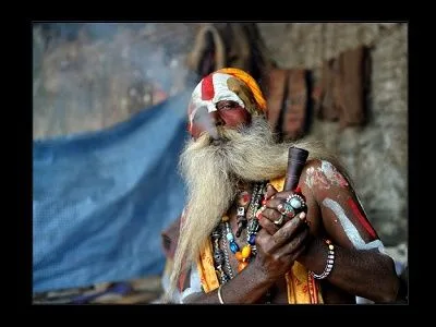 Hombre de larga barba blanca y rostro pintado sostiene una pipa entre las manos, con humo alrededor y una tela azul al fondo.