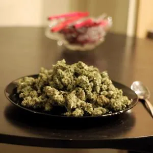 Plato negro lleno de cogollos de cannabis sobre una mesa oscura, con una cuchara al lado y un recipiente desenfocado al fondo.