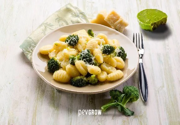 Plato de ñoquis con brócoli y queso rallado, acompañado de trozos de queso, una porción verde y un tenedor sobre una mesa blanca. Plato de ñoquis con brócoli y queso rallado, acompañado de trozos de queso, una porción verde y un tenedor sobre una mesa blanca.
