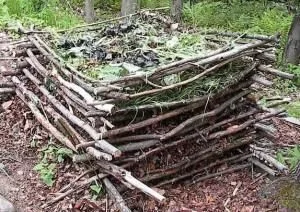 Compostera cuadrada construida con ramas apiladas y entrecruzadas, llena de restos vegetales en un entorno arbolado. Compostera cuadrada construida con ramas apiladas y entrecruzadas, llena de restos vegetales en un entorno arbolado.