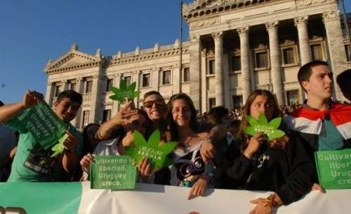 Concentración ante un edificio de columnas, con carteles verdes, siluetas de hojas de cannabis y una bandera uruguaya sobre los hombros de un participante. Concentración ante un edificio de columnas, con carteles verdes, siluetas de hojas de cannabis y una bandera uruguaya sobre los hombros de un participante.