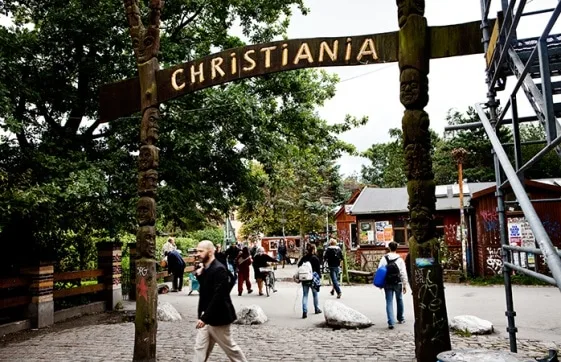 Entrada con un arco de madera rotulado CHRISTIANIA, varias personas en el camino, árboles y edificios al fondo. Entrada con un arco de madera rotulado CHRISTIANIA, varias personas en el camino, árboles y edificios al fondo.