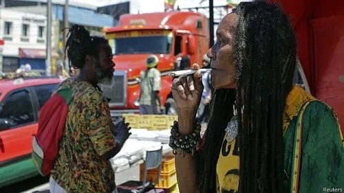 Persona con rastas fumando un cigarrillo enrollado en primer plano, con otra persona, coches y un camión rojo detrás. Persona con rastas fumando un cigarrillo enrollado en primer plano, con otra persona, coches y un camión rojo detrás.