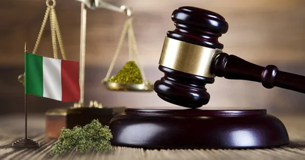 Composición con un mazo judicial sobre su base, una bandera italiana, una balanza y cogollos de cannabis en primer plano. Composición con un mazo judicial sobre su base, una bandera italiana, una balanza y cogollos de cannabis en primer plano.