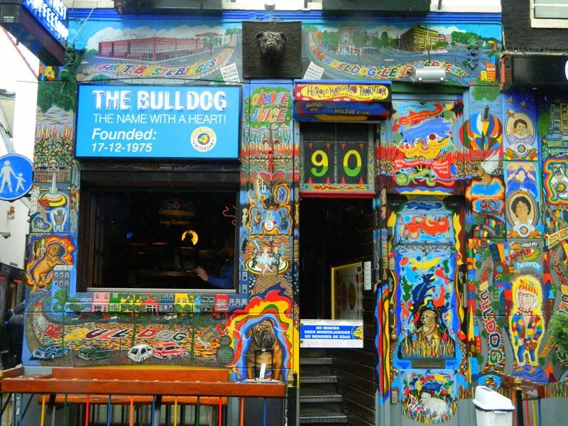 Fachada de The Bulldog cubierta de murales multicolores, con cartel azul, número 90 sobre la entrada y un banco delante. Fachada de The Bulldog cubierta de murales multicolores, con cartel azul, número 90 sobre la entrada y un banco delante.