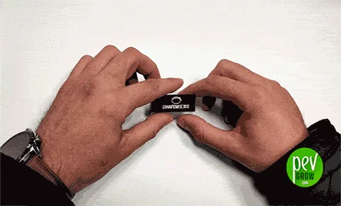 Dos manos sujetan una pequeña pieza rectangular negra con un logotipo blanco sobre una mesa blanca; la muñeca izquierda lleva reloj.
