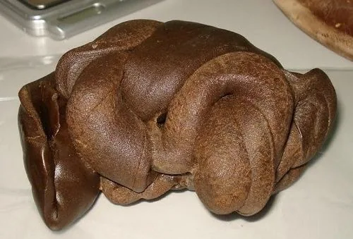 Masa de hachís marrón compacto con superficie lisa, pliegues y formas redondeadas, apoyada sobre una superficie clara. Masa de hachís marrón compacto con superficie lisa, pliegues y formas redondeadas, apoyada sobre una superficie clara.