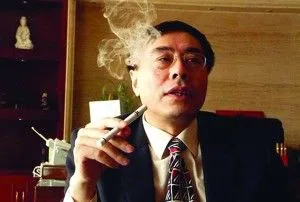 Hombre con gafas, traje y corbata sostiene un dispositivo alargado cerca de la boca, con una nube de humo o vapor ante el rostro.