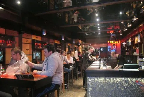 Interior de un coffeeshop con clientes en mesas altas, una barra iluminada y un techo oscuro con focos. Interior de un coffeeshop con clientes en mesas altas, una barra iluminada y un techo oscuro con focos.