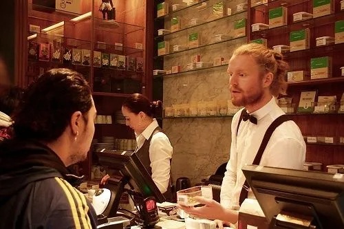Un empleado con pajarita y tirantes atiende a un cliente ante un mostrador, con estantes de cajas al fondo. Un empleado con pajarita y tirantes atiende a un cliente ante un mostrador, con estantes de cajas al fondo.