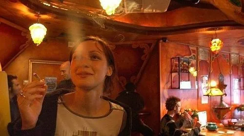 Mujer en primer plano sostiene un cigarrillo encendido en un local de tonos rojizos, con lámparas colgantes y otras personas al fondo. Mujer en primer plano sostiene un cigarrillo encendido en un local de tonos rojizos, con lámparas colgantes y otras personas al fondo.