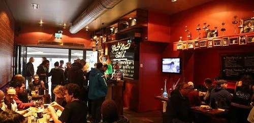 Interior de un coffeeshop con paredes rojas, clientes en mesas y junto a la barra, una pantalla y trofeos en una repisa. Interior de un coffeeshop con paredes rojas, clientes en mesas y junto a la barra, una pantalla y trofeos en una repisa.