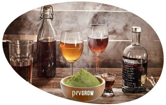 Composición ovalada con un cuenco de polvo verde, botellas, copas y vasos con bebidas de distintos tonos sobre una mesa de madera.