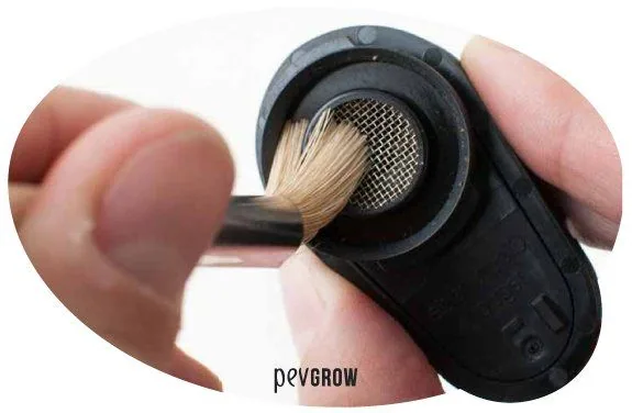 Primer plano de unas manos que limpian con un pincel la rejilla metálica circular de un vaporizador negro.