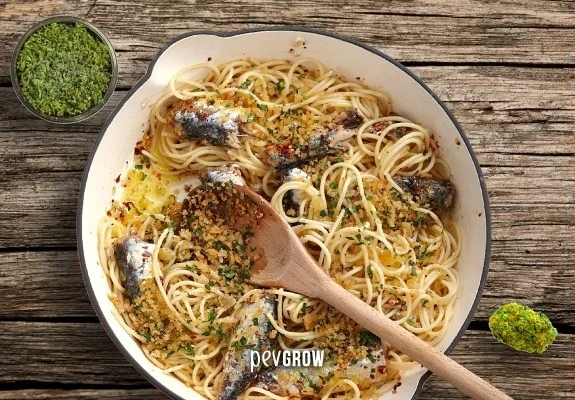 Espaguetis con sardinas, hierbas y migas en una sartén clara, con una cuchara de madera y un cuenco de hierbas al lado. Espaguetis con sardinas, hierbas y migas en una sartén clara, con una cuchara de madera y un cuenco de hierbas al lado.