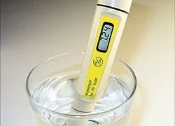 Medidor digital de pH amarillo con la sonda sumergida en un vaso transparente con agua, visto en primer plano.