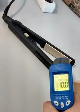 Termómetro infrarrojo azul apuntando a la placa de una plancha de pelo abierta, con una lectura de 110,0 °C en pantalla. Termómetro infrarrojo azul apuntando a la placa de una plancha de pelo abierta, con una lectura de 110,0 °C en pantalla.