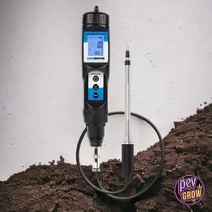 Medidor digital negro con pantalla azul, cable y sonda alargada, colocado en vertical junto a un montículo de tierra. Medidor digital negro con pantalla azul, cable y sonda alargada, colocado en vertical junto a un montículo de tierra.