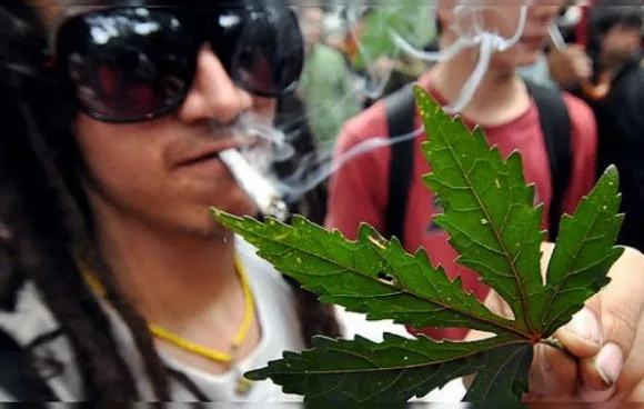 Persona con gafas de sol y un cigarrillo entre los labios sostiene una hoja de cannabis en primer plano, con humo alrededor. Persona con gafas de sol y un cigarrillo entre los labios sostiene una hoja de cannabis en primer plano, con humo alrededor.