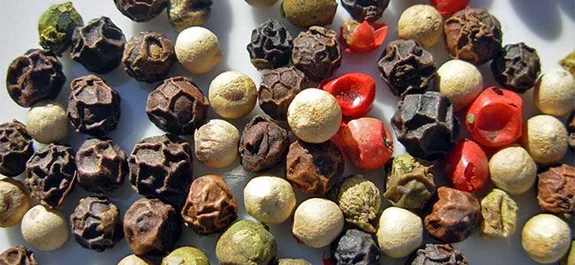 Primer plano de granos de pimienta negros, blancos, verdes y rojos, dispersos sobre una superficie clara.