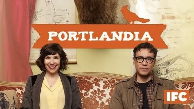 Una mujer sonriente y un hombre con gafas sentados en un sofá estampado, bajo el rótulo naranja de Portlandia.