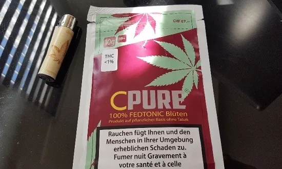 Envase CPURE con una hoja de cannabis, indicaciones de 10 g y THC inferior al 1 %, junto a un mechero sobre una mesa oscura. Envase CPURE con una hoja de cannabis, indicaciones de 10 g y THC inferior al 1 %, junto a un mechero sobre una mesa oscura.