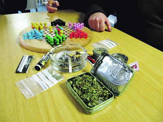 Mesa con una lata abierta de cannabis, un cenicero, un cigarrillo liado y un tablero de fichas de colores junto a varias personas.