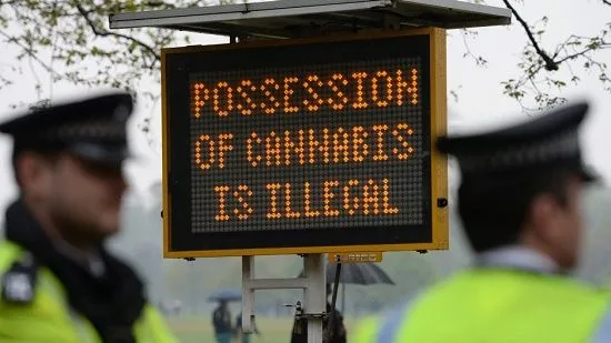 Panel luminoso con el mensaje en inglés «La posesión de cannabis es ilegal», entre dos agentes desenfocados. Panel luminoso con el mensaje en inglés «La posesión de cannabis es ilegal», entre dos agentes desenfocados.