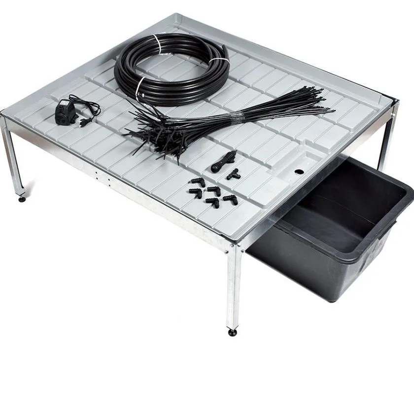 Mesa de cultivo con bandeja acanalada, rollo de tubo negro, tubos finos, bomba y conectores; un depósito negro sobresale por debajo.