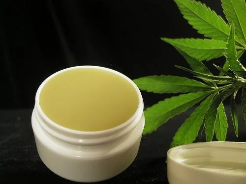 Tarro blanco abierto con crema amarillenta, junto a su tapa y hojas de cannabis sobre una superficie oscura. Tarro blanco abierto con crema amarillenta, junto a su tapa y hojas de cannabis sobre una superficie oscura.