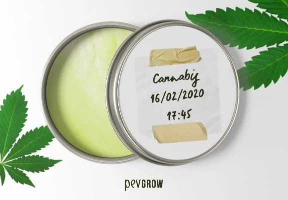 Lata abierta con crema amarillenta y tapa con etiqueta manuscrita, rodeada de hojas de cannabis sobre fondo blanco. Lata abierta con crema amarillenta y tapa con etiqueta manuscrita, rodeada de hojas de cannabis sobre fondo blanco.