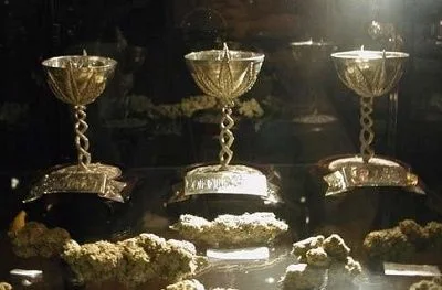Tres trofeos dorados con pies en espiral, expuestos tras un cristal junto a varios cogollos de cannabis.