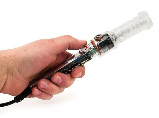 Una mano sostiene en diagonal un vaporizador alargado con cuerpo transparente, componentes internos visibles y un cable negro sobre fondo blanco.